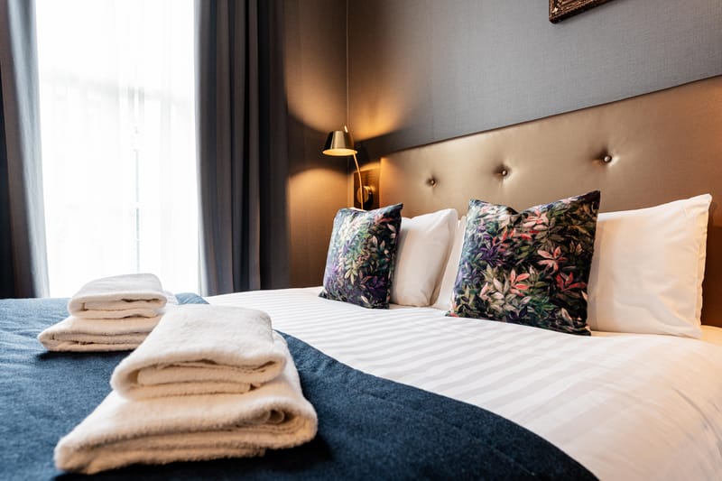 A double bed by the window, in white linen with a navy runner and botanical cushions
