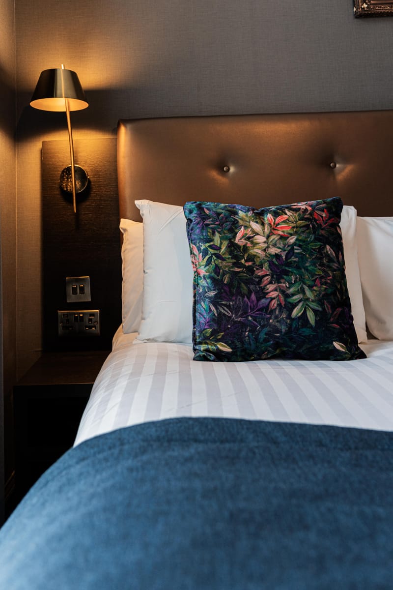 A single bed in white linen with a navy runner and a botanical velvet cushion