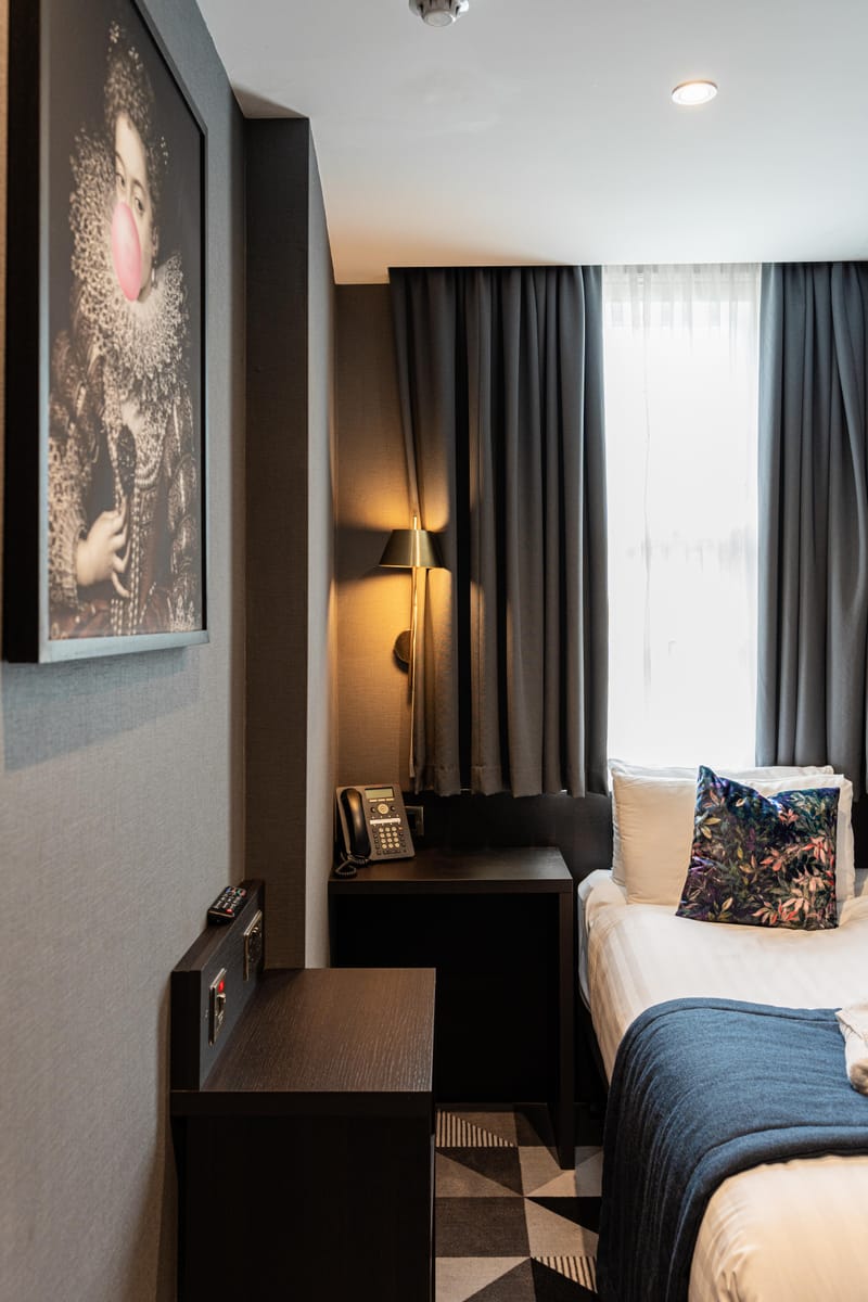 A single room with a bed dressed in white linen and a navy runner beneath a framed portrait